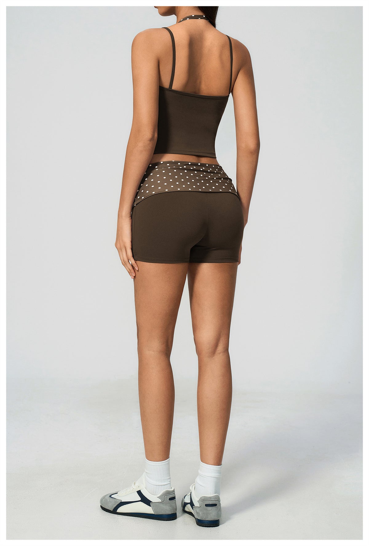 A model shown from the back wearing a dark brown activewear set consisting of a spaghetti-strap crop top and matching fitted shorts with a sheer mesh back panel and silver studs. The model is also wearing white socks and gray athletic sandals against a light gray studio background.