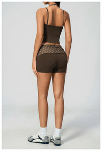 A model shown from the back wearing a dark brown activewear set consisting of a spaghetti-strap crop top and matching fitted shorts with a sheer mesh back panel and silver studs. The model is also wearing white socks and gray athletic sandals against a light gray studio background.