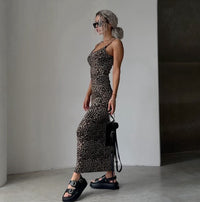 A woman wearing a sleeveless leopard-print maxi dress, black platform sandals, and sunglasses, holding a small black handbag in a minimalist indoor setting.