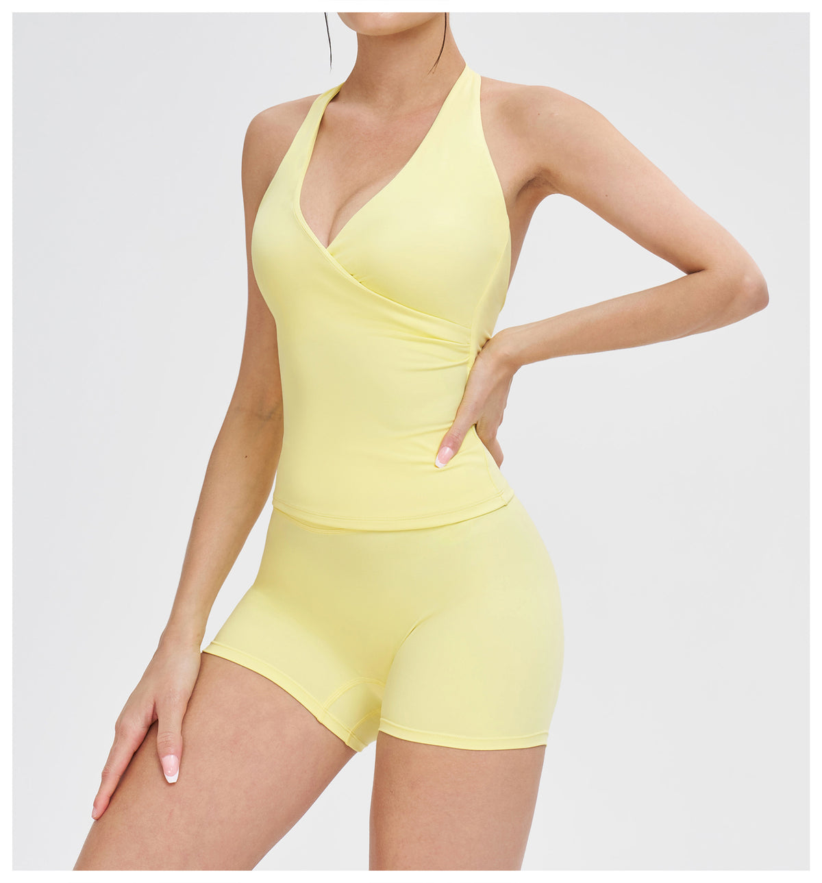 A woman modeling a pale yellow one-piece athletic romper with a wrap-style V-neck bodice and fitted shorts against a light gray studio background.