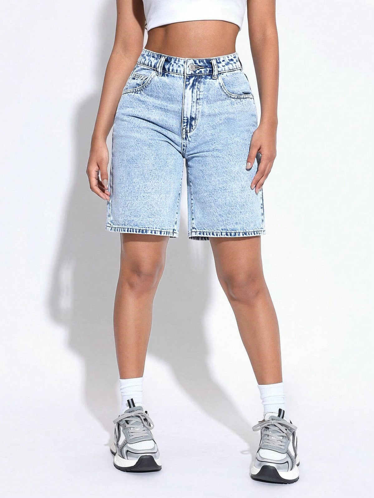 A model wearing light-wash blue denim shorts with a raw hem, styled with white crew socks and gray sneakers against a white studio background.