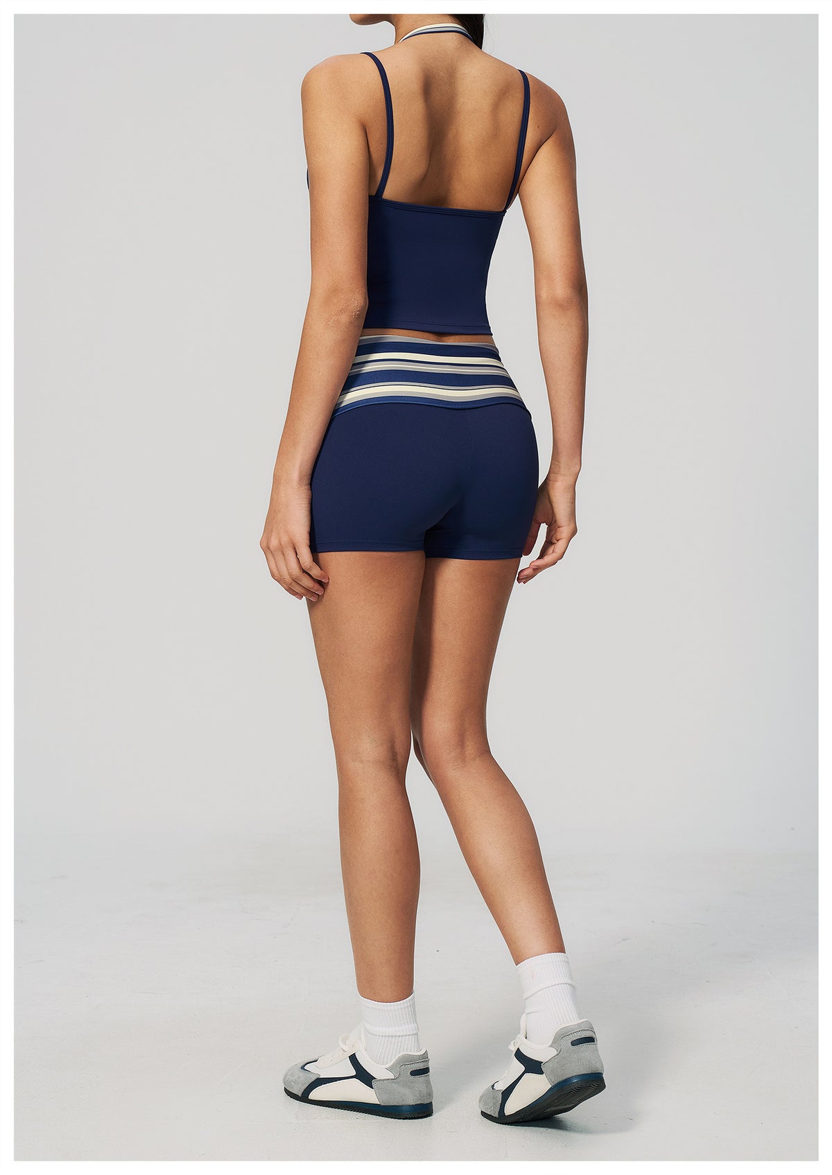 A model shown from the back wearing a navy blue activewear set consisting of a spaghetti-strap crop top and matching high-waisted shorts with white and light blue horizontal stripes at the waistband, paired with white socks and gray athletic sandals on a light gray background.