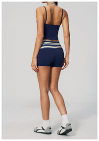 A model shown from the back wearing a navy blue activewear set consisting of a spaghetti-strap crop top and matching high-waisted shorts with white and light blue horizontal stripes at the waistband, paired with white socks and gray athletic sandals on a light gray background.