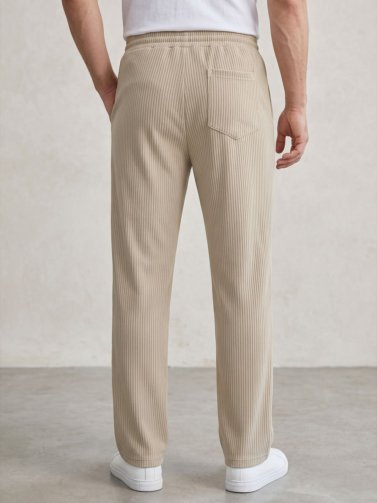 A person shown from the back wearing beige ribbed trousers with a back pocket, paired with a white shirt and white sneakers, against a neutral studio backdrop.