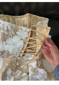A close-up of a beige and white floral lace corset dress with a hand adjusting the lace-up closure, set against a black mesh background.