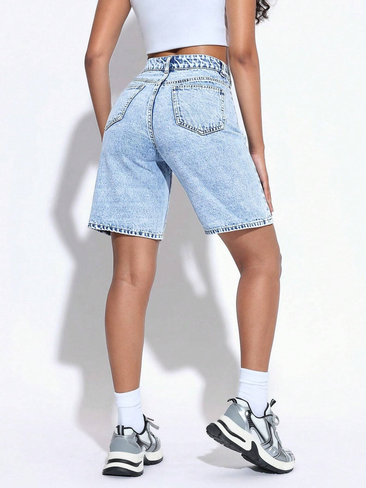 A person shown from the back wearing light-wash denim shorts, a white cropped top, white socks, and gray sneakers against a plain white studio background.