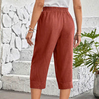 A person wearing rust-colored cropped pants with an elastic waistband, standing in front of a white stone wall and green plant.