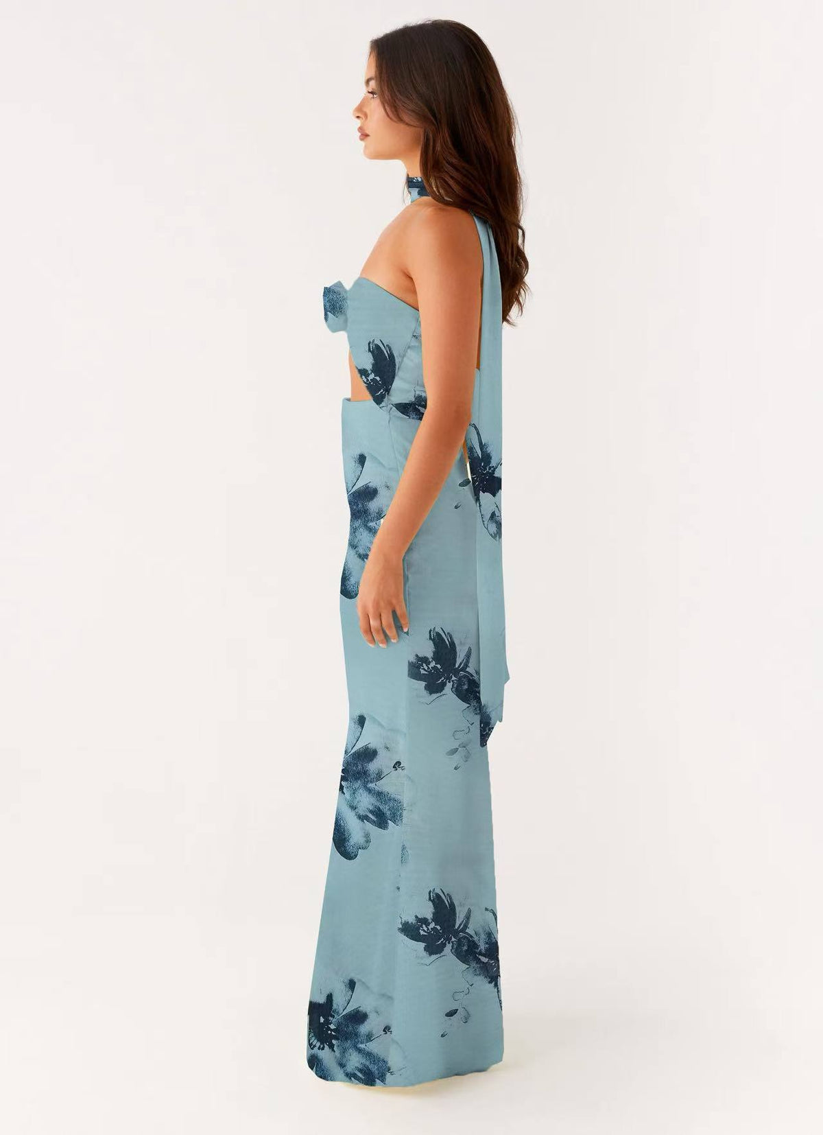 A woman modeling a light blue halter-neck maxi dress with a dark blue floral print, shown from a side profile against a plain white studio background.