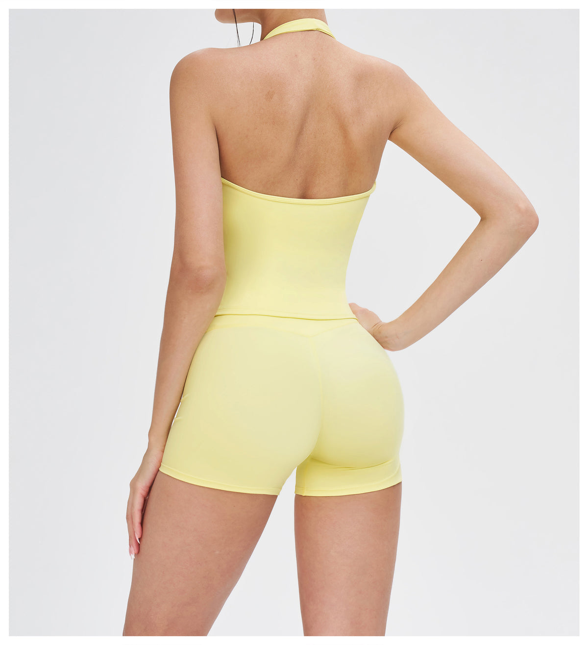 A model shown from the back wearing a pale yellow halter-neck bodysuit with matching high-waisted shorts against a light gray studio background.