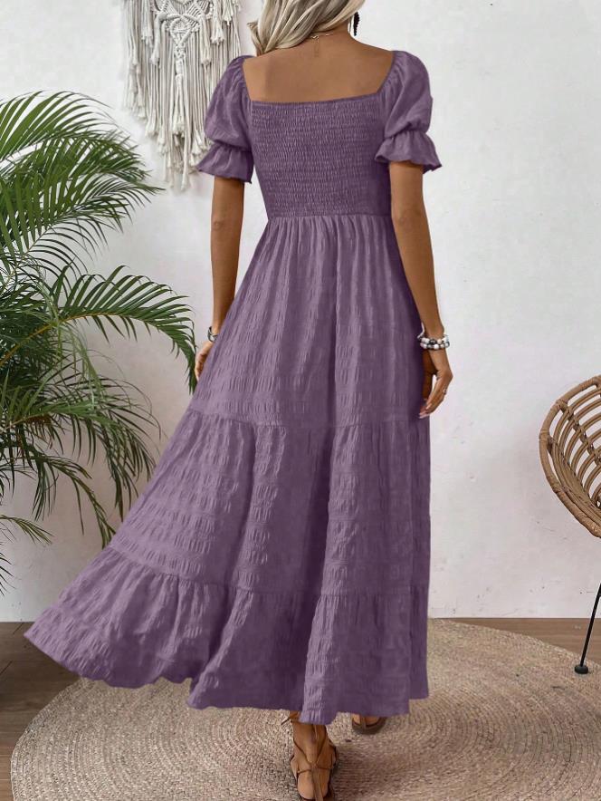 A woman wearing a lavender purple tiered maxi dress with puff sleeves and a smocked bodice, shown from the back in a styled indoor setting with a plant and woven chair.