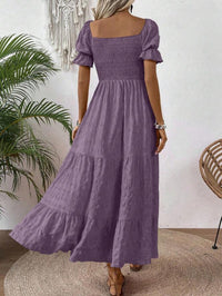 A woman wearing a lavender purple tiered maxi dress with puff sleeves and a smocked bodice, shown from the back in a styled indoor setting with a plant and woven chair.