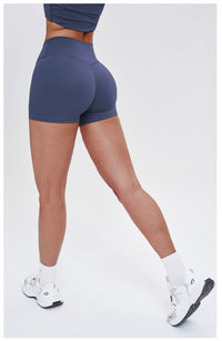 Rear view of a model wearing high-waisted, fitted athletic shorts in a muted slate blue color, paired with white socks and white sneakers, against a plain light gray studio background.