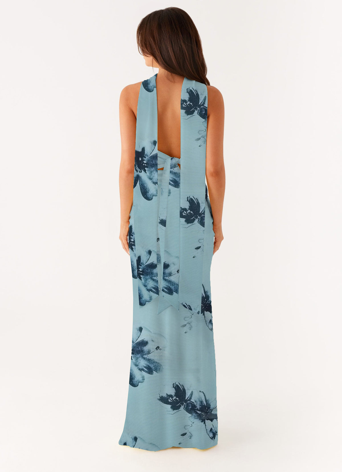 A woman viewed from the back wearing a sleeveless, floor-length halter-style dress with a light blue base and dark navy floral print, against a plain light background.