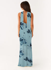 A woman viewed from the back wearing a sleeveless, floor-length halter-style dress with a light blue base and dark navy floral print, against a plain light background.