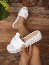 White espadrille-style slide sandals with a wide decorative strap and braided jute-wrapped platform soles, shown on a wooden floor with one sandal worn and the other held in hand.