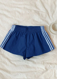 Blue athletic shorts with white three-stripes on the sides, laid flat on a textured off-white fabric background with a small white cup in the top right corner.