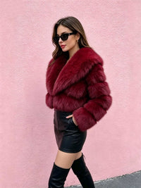 A woman wearing a voluminous deep burgundy faux fur jacket, black leather mini skirt, and black thigh-high boots, posing against a textured light pink wall.