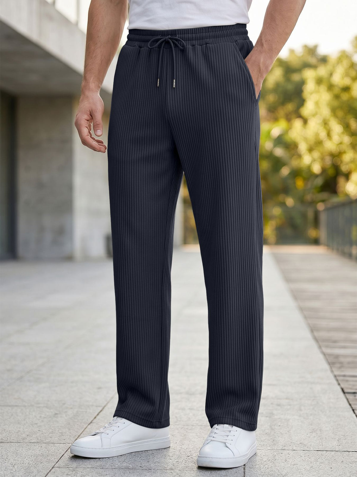 A person wearing dark charcoal gray pinstriped drawstring trousers, styled with a white t-shirt and white sneakers, standing on a paved outdoor walkway.