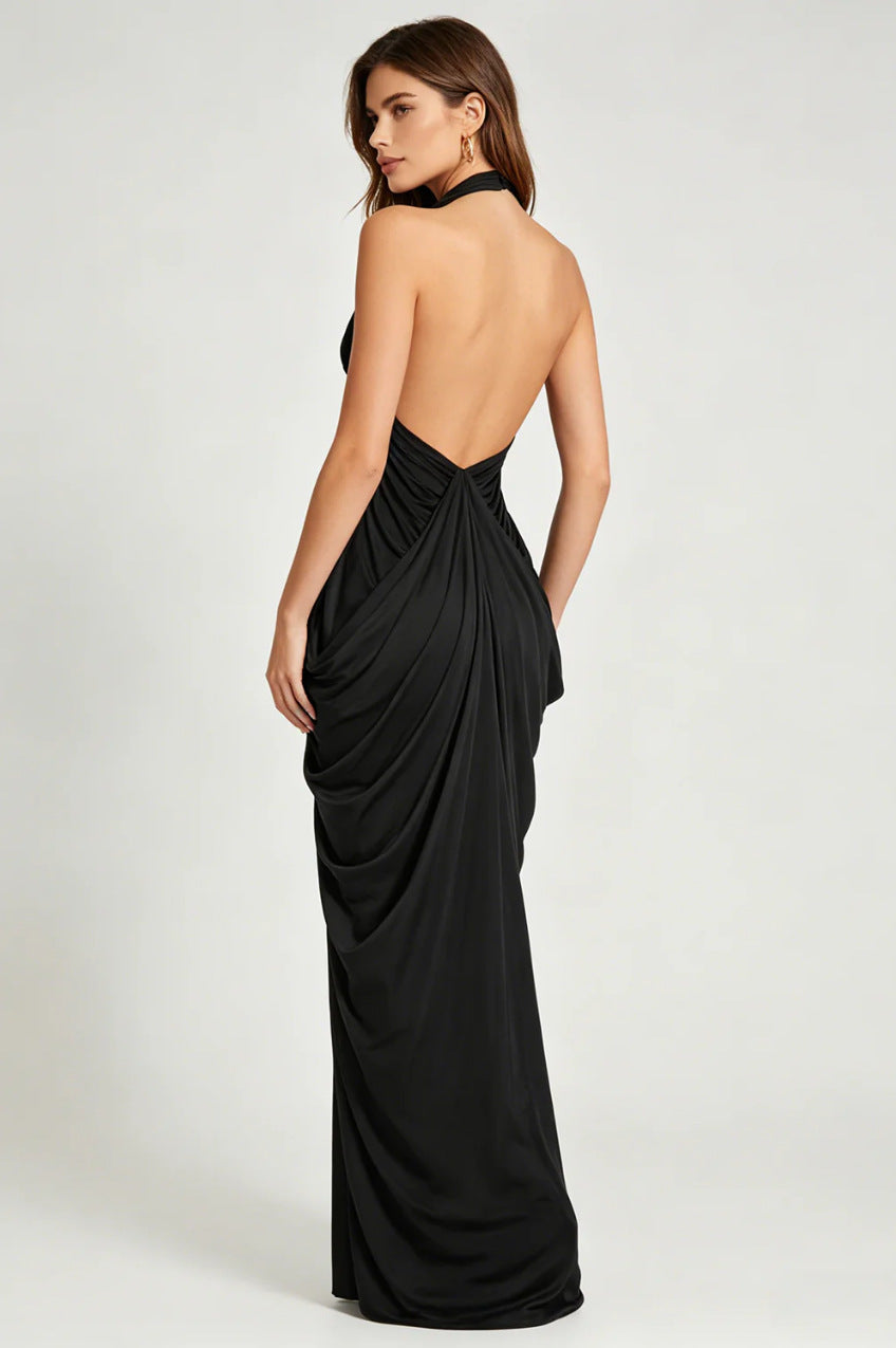 A woman modeling a floor-length black halter gown from the back, showing a draped open back and gathered waist, against a plain light gray studio background.