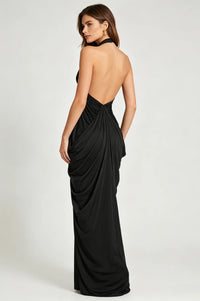 A woman modeling a floor-length black halter gown from the back, showing a draped open back and gathered waist, against a plain light gray studio background.