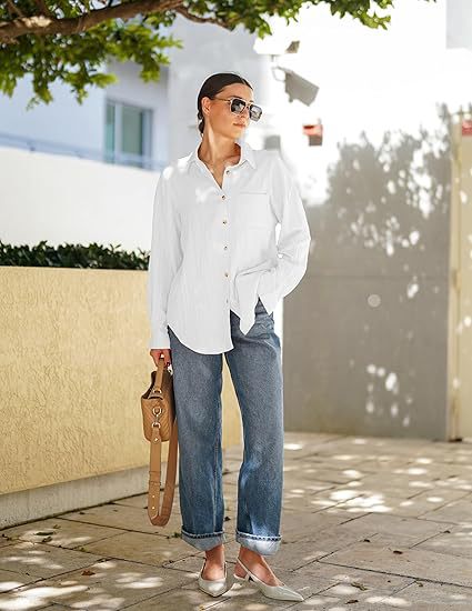 A woman stands outdoors wearing a white long-sleeve button-up shirt, blue jeans, and light-colored pointed shoes, holding a tan shoulder bag. The background shows a sunlit urban walkway with a beige wall, tree shadows, and a building.
