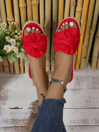 Red open-toe wedge sandals with large fabric bows on the toes, shown worn with blue jeans and a beaded ankle bracelet against a bamboo and wood floor background.