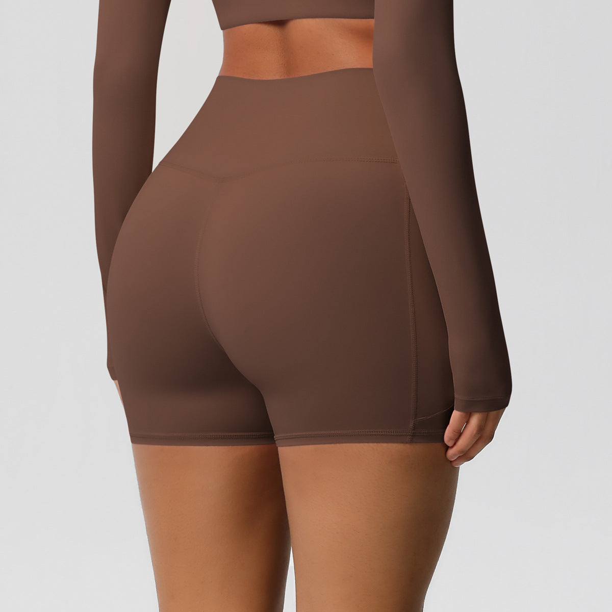 Rear view of a model wearing high-waisted, long-sleeve athletic shorts in a solid medium brown color, paired with a matching long-sleeve crop top, against a light gray studio background.