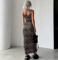 A woman stands with her back to the camera wearing a fitted leopard-print maxi dress with thin straps and a low open back. She is holding a small black handbag and wearing black shoes in a minimalist indoor setting with white walls and a concrete floor.