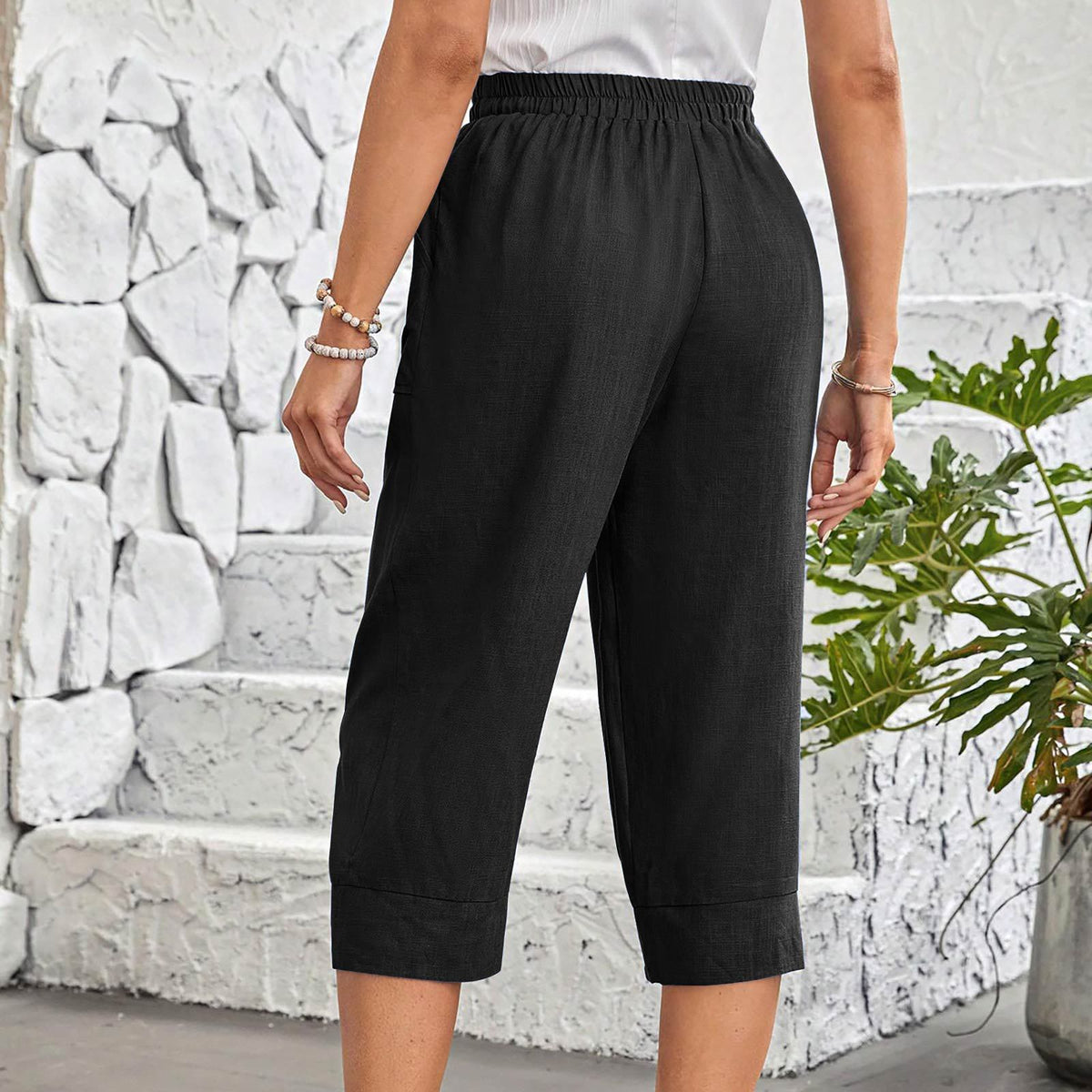A person wearing black cropped pants with an elastic waistband and rolled cuffs, shown from the back in an outdoor setting with a white stone wall and green plant.