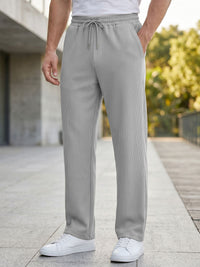 A person wearing light gray pinstriped drawstring lounge pants, styled with a white t-shirt and white sneakers, standing on a paved outdoor walkway.