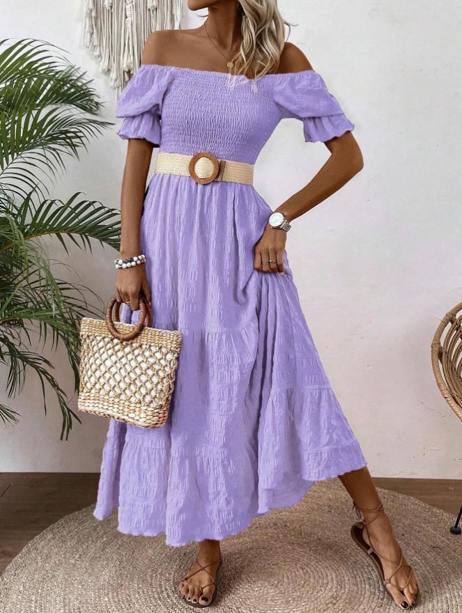 A woman models an off-the-shoulder lavender midi dress with a smocked bodice, tiered skirt, and a beige belt with a round wooden buckle. She is holding a woven straw handbag and wearing flat brown sandals, standing on a round jute rug against a white wall with a plant in the background.