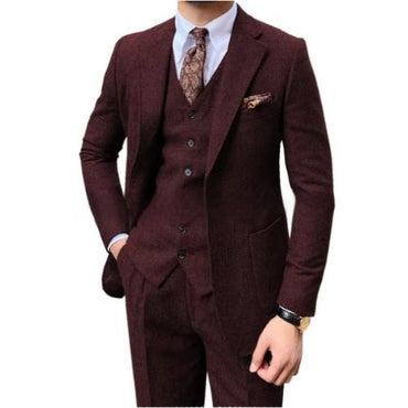 A men's three-piece suit in a deep burgundy or maroon textured fabric, featuring a jacket, matching vest, and trousers, styled with a white dress shirt, a brown paisley tie, and a matching pocket square on a white background.