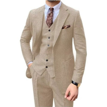 A man wearing a beige three-piece suit with a matching vest and trousers, styled with a white dress shirt, a brown patterned tie, and a matching pocket square, against a plain white background.