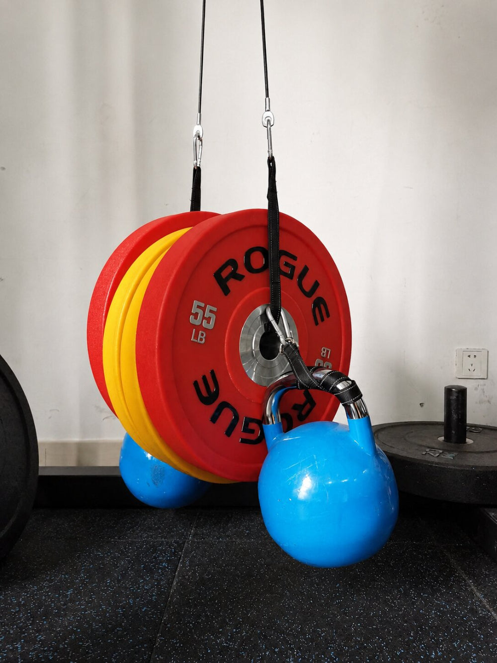 A blue kettlebell hanging from a black cable system attached to red and yellow Rogue 55 lb weight plates in a gym setting.
