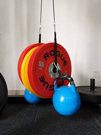 A blue kettlebell hanging from a black cable system attached to red and yellow Rogue 55 lb weight plates in a gym setting.
